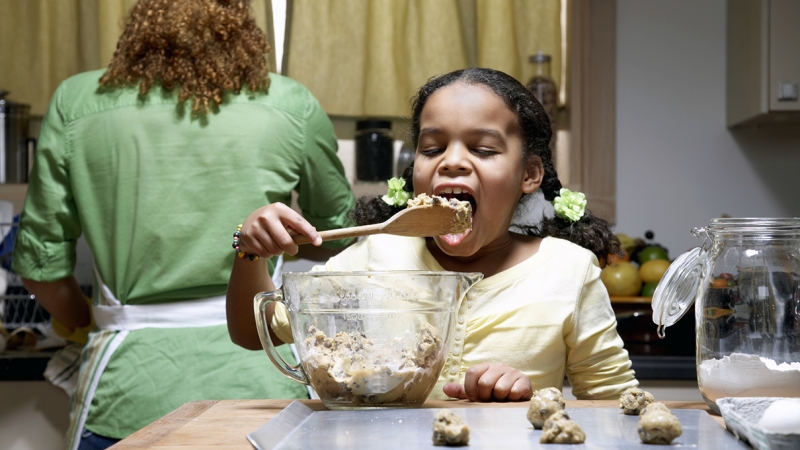 Girl (8-10) licking spoon from dough mixture