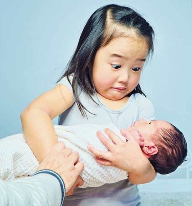 Vẻ mặt hài hước của bé gái khi lần đầu ẵm em trai, nặng hơn hẳn so với con búp bê em chơi thường ngày.