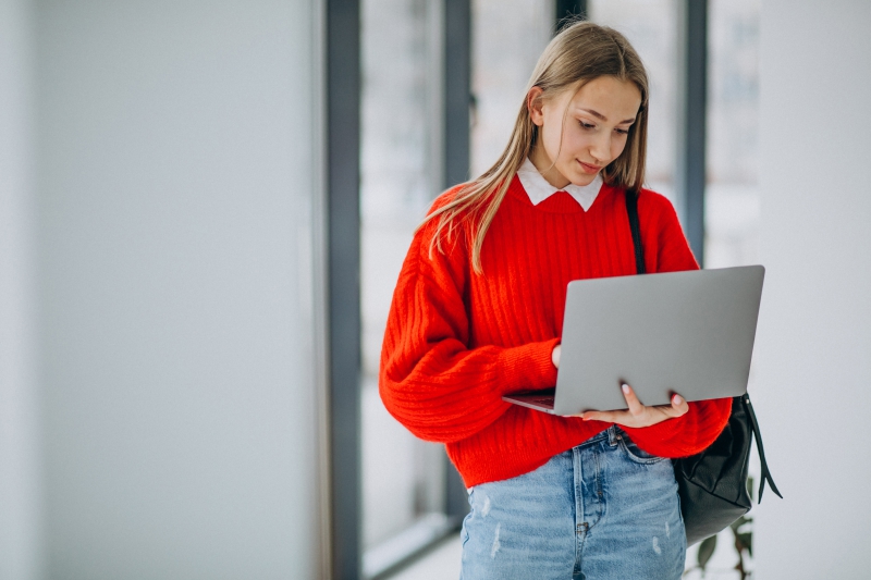 Girl student with laptop standing by the window in corridor